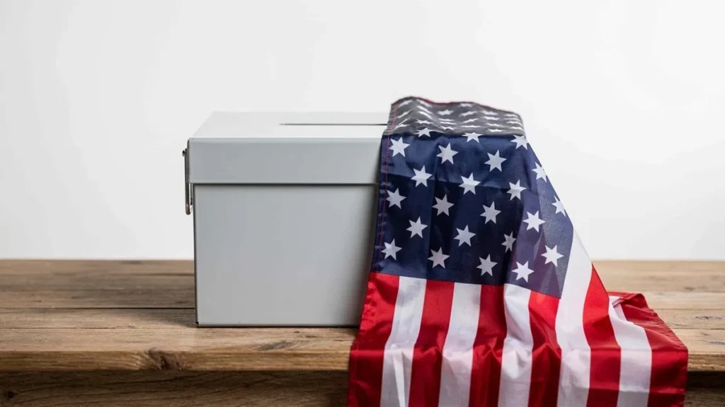 White ballot box draped with an American flag on a wooden table